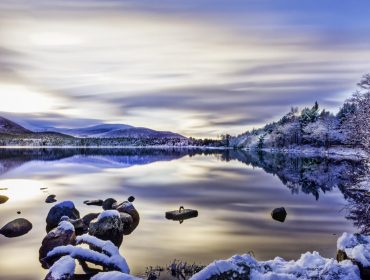 Beautiful winters day with soft clouds, snow on trees and rocks, reflections on calm water at Loch Morlich, Aviemore